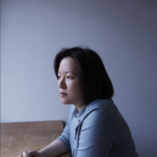 A woman sits at a table. She has shoulder-length, black hair and wears a light blue shirt.