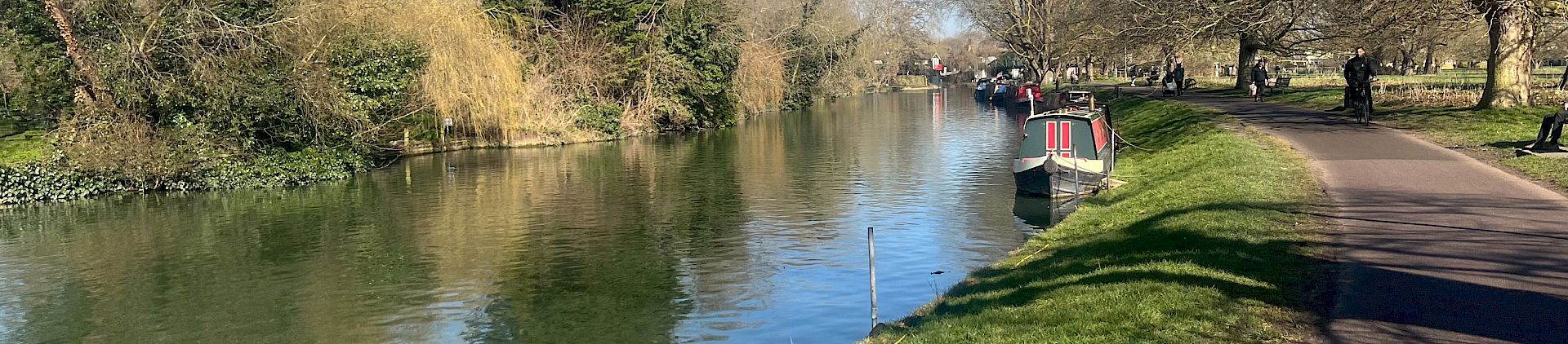 A photograph of the river Cam. A grassy bank in the foreground and a canal boat in the distance. Blue sky overhead.
