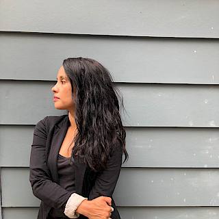 Woman with long dark hair stands against a wooden slatted wall. She wears jeans and a black blazer.