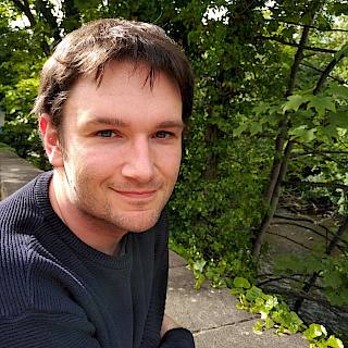 Photo of a white man with short brown hair, wearing a navy blue jumper. In the background are some trees.
