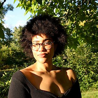 A woman with an afro, glasses, and a black top stands facing the camera. The background shows grass and trees.