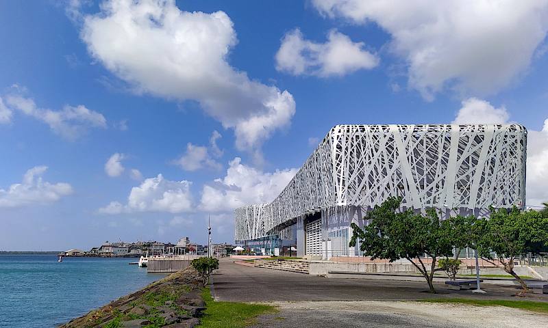 A silver metallic building sits against a blue sky. The sea is to the left.