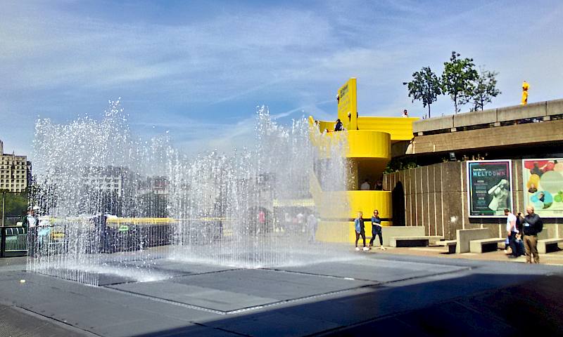 On the right is a concrete building with a bright yellow circular staircase. On the left are some waterfountains shooting into the air.