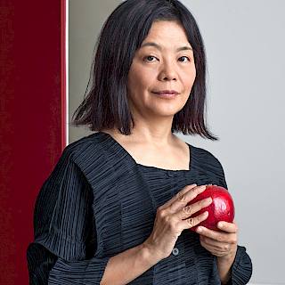 Woman with shoulder length straight black hair wearing a navy blue long-sleeved top stands holding a red apple.