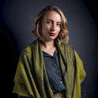 Photo of a woman shown from the waist up. She has shoulder-length blond curly hair and red lipstick. She wears a blue shirt with a green shawl and beige trousers. She stands against a black backdrop.