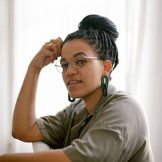 Photo of a person sat with their arm on the back of a chair. Their head is resting on their fist. They have black braids and large green earrings. They have circular frameless glasses and wear a grey top.
