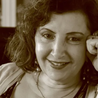 Sepia photo of a person with shoulder length curly hair. They are resting their head on their fist and are wearing a dark coloured vest and a light cardigan.