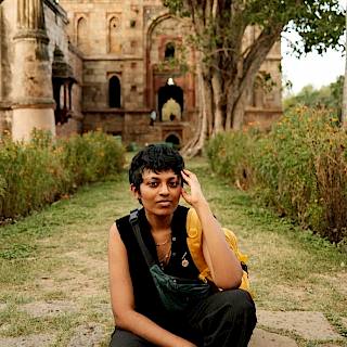 Photo of a person in a yellow top and black dungarees sat on a step with an ancient monument and greenery behind them. They have short dark hair and are resting their head on their hand.
