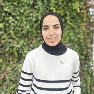A woman in a black hijab sends against a hedge. She is wearing a black and white striped long sleeved top.