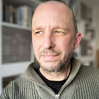 A bald man looks off to the side. He is wearing a green zip up jumper and there is a bookcase behind him.