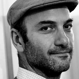 Black and white photo of a man with light stubble wearing a flat cap and jumper over a checked shirt.