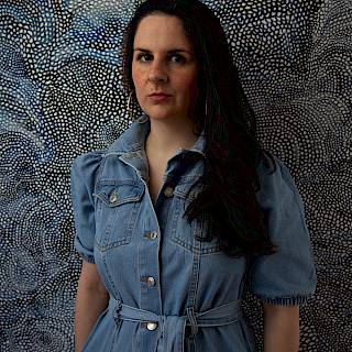 A photo of a woman shown from the hips up. She has long dark hair and is wearing a blue denim dress. She stands against blue patterned wallpaper.