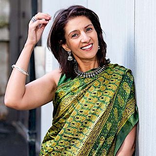 Woman in a green and yellow sari stands leaning against a white wall. She is smiling and has her hand raised towards her hair.