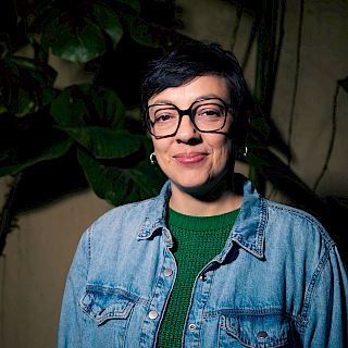 Photo of a woman with short black hair. She wears black framed glasses and gold earrings. She wears a green top with a denim shirt over the top.